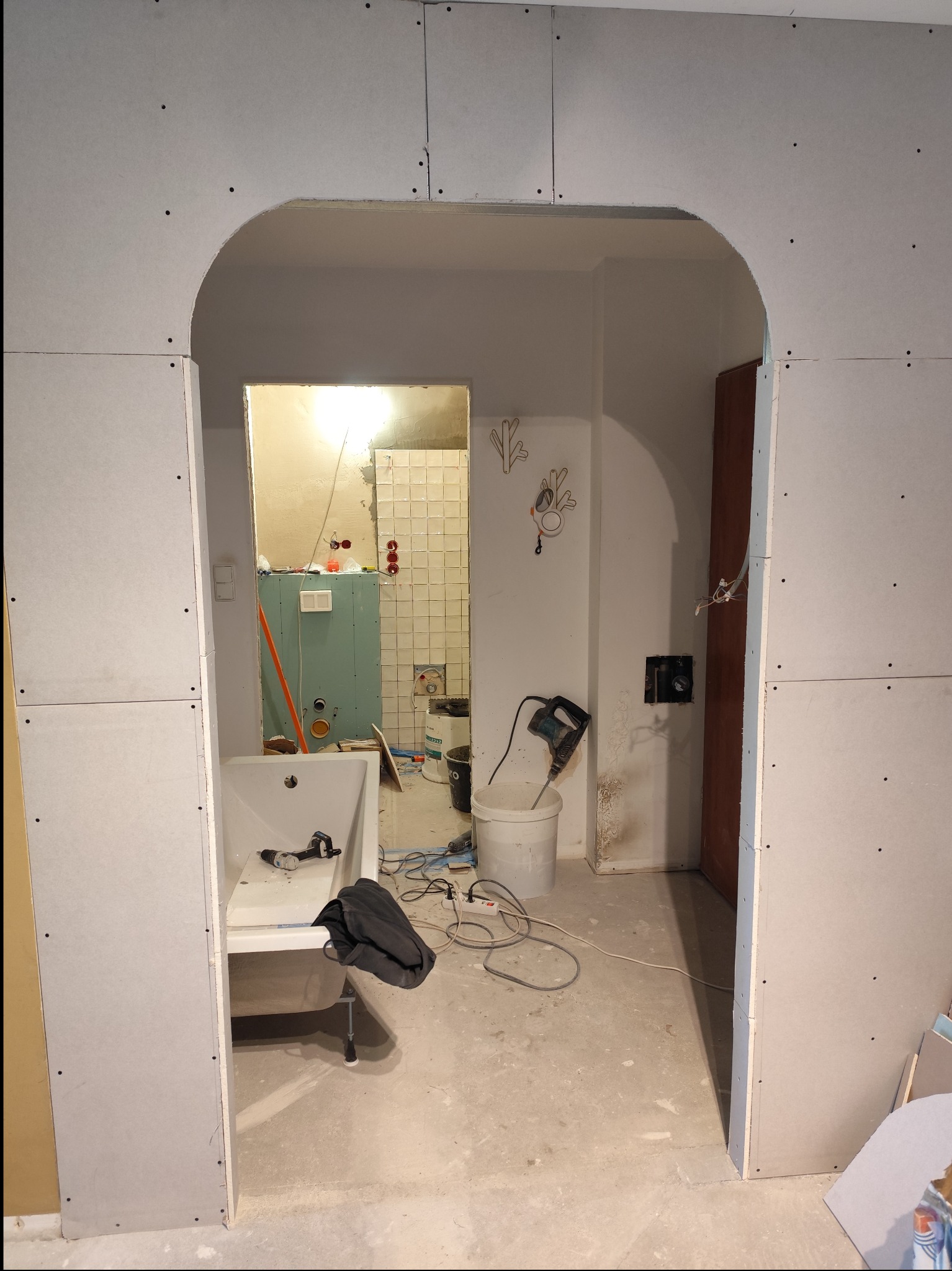 Łazienka w trakcie remontu: widoczna wanna, płyty gipsowo-kartonowe na ścianach z wyciętym łukiem, narzędzia i akcesoria budowlane porozrzucane na podłodze, fragment wykafelkowanej ściany w głębi...