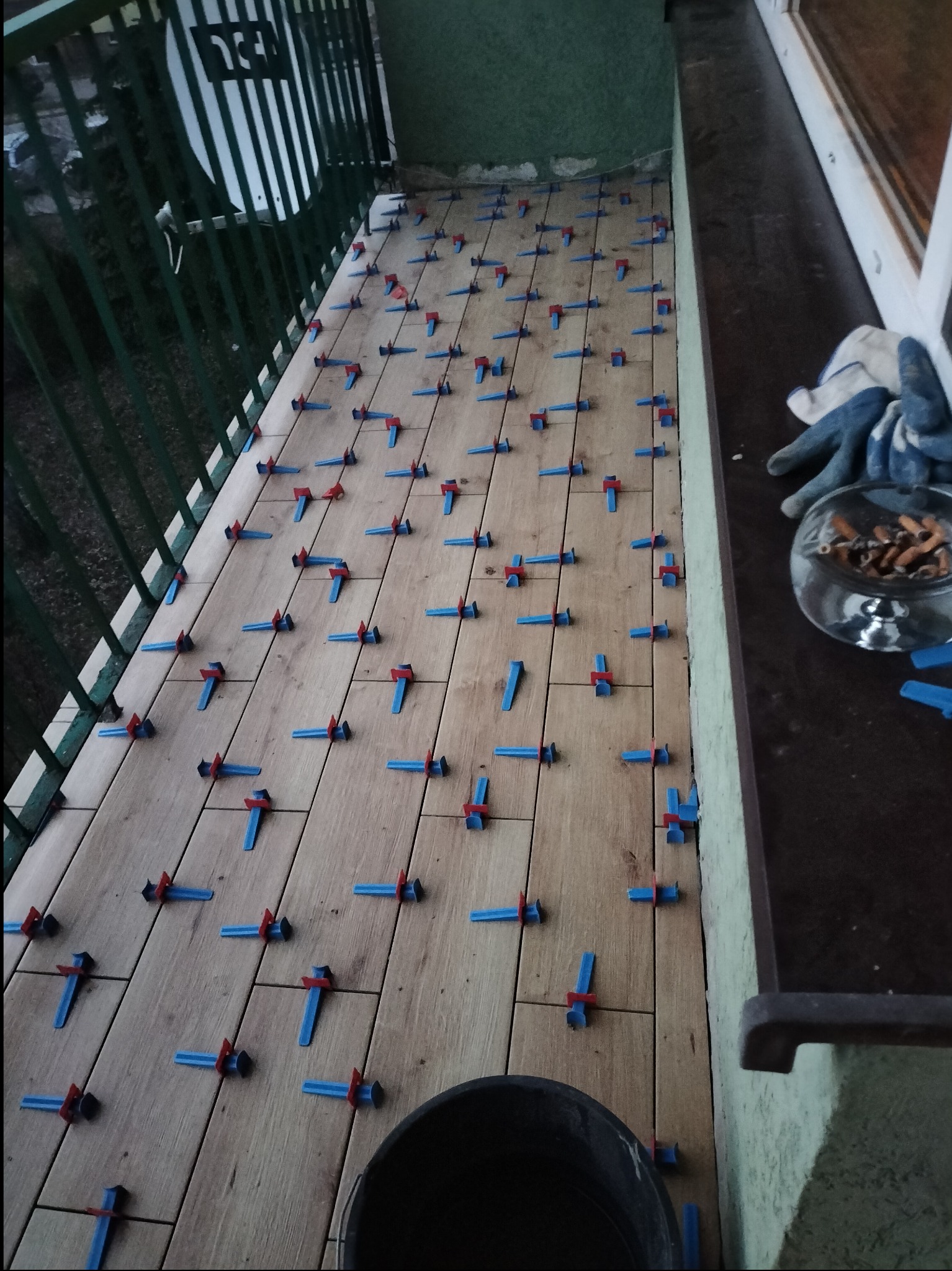 Układanie płytek drewnopodobnych na balkonie z użyciem niebiesko-czerwonych klipsów poziomujących, widoczny fragment wiadra i popielniczka z niedopałkami.