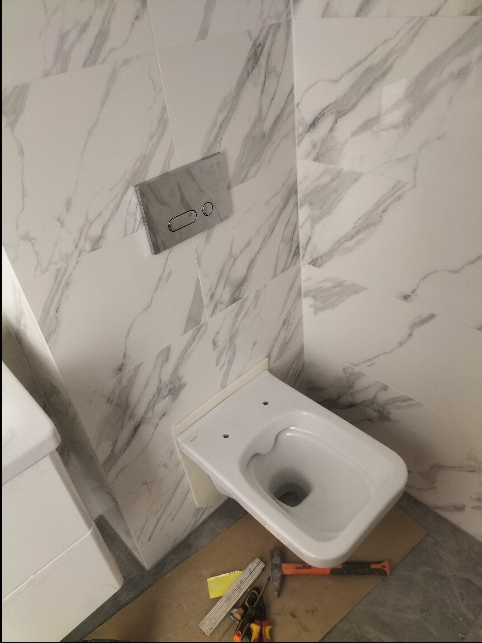 Wisząca miska WC w trakcie montażu na tle ściany wyłożonej płytkami imitującymi marmur, widoczne narzędzia na podłodze.