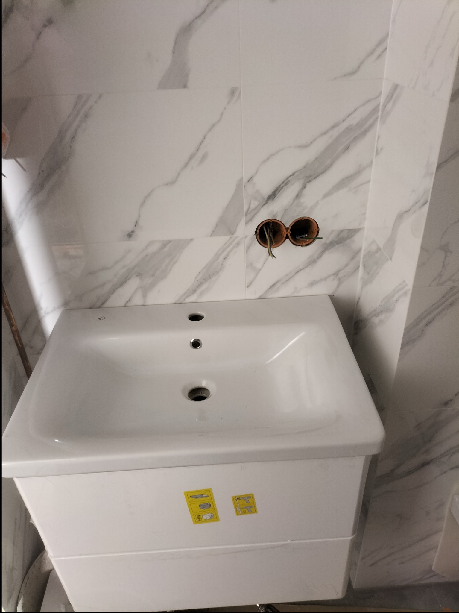 Biała umywalka z szafką zamontowana na tle ściany wyłożonej płytkami imitującymi marmur, widoczne puszki elektryczne z wystającymi przewodami.