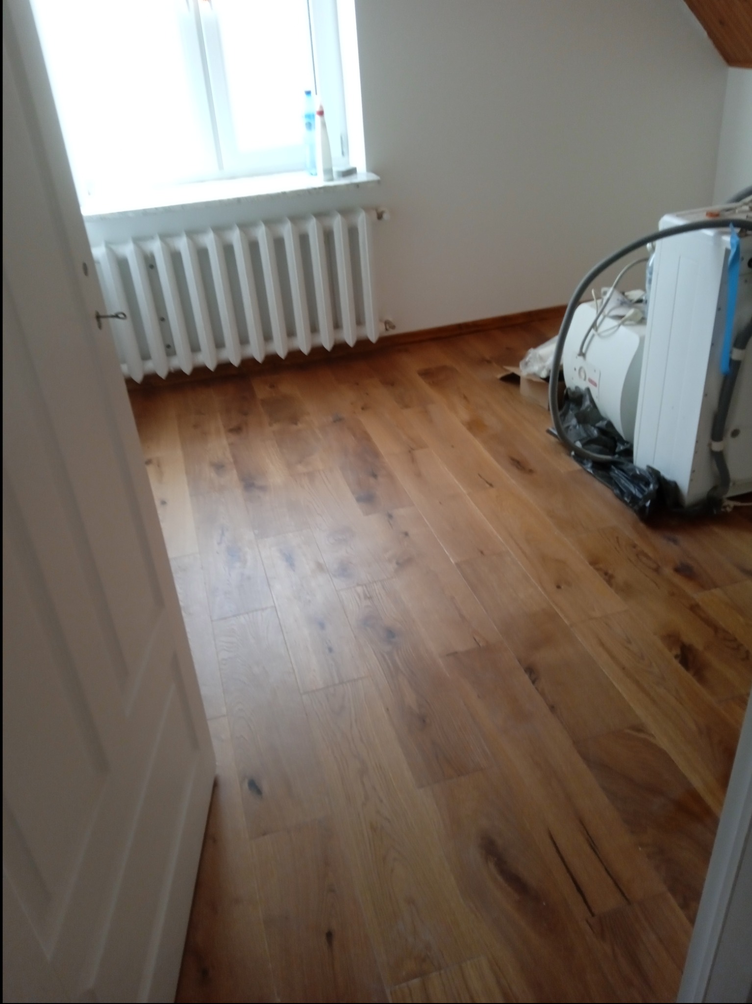Ukończona podłoga z paneli drewnianych w jasnym odcieniu, widoczny grzejnik pod oknem i sprzęt budowlany w rogu pokoju.