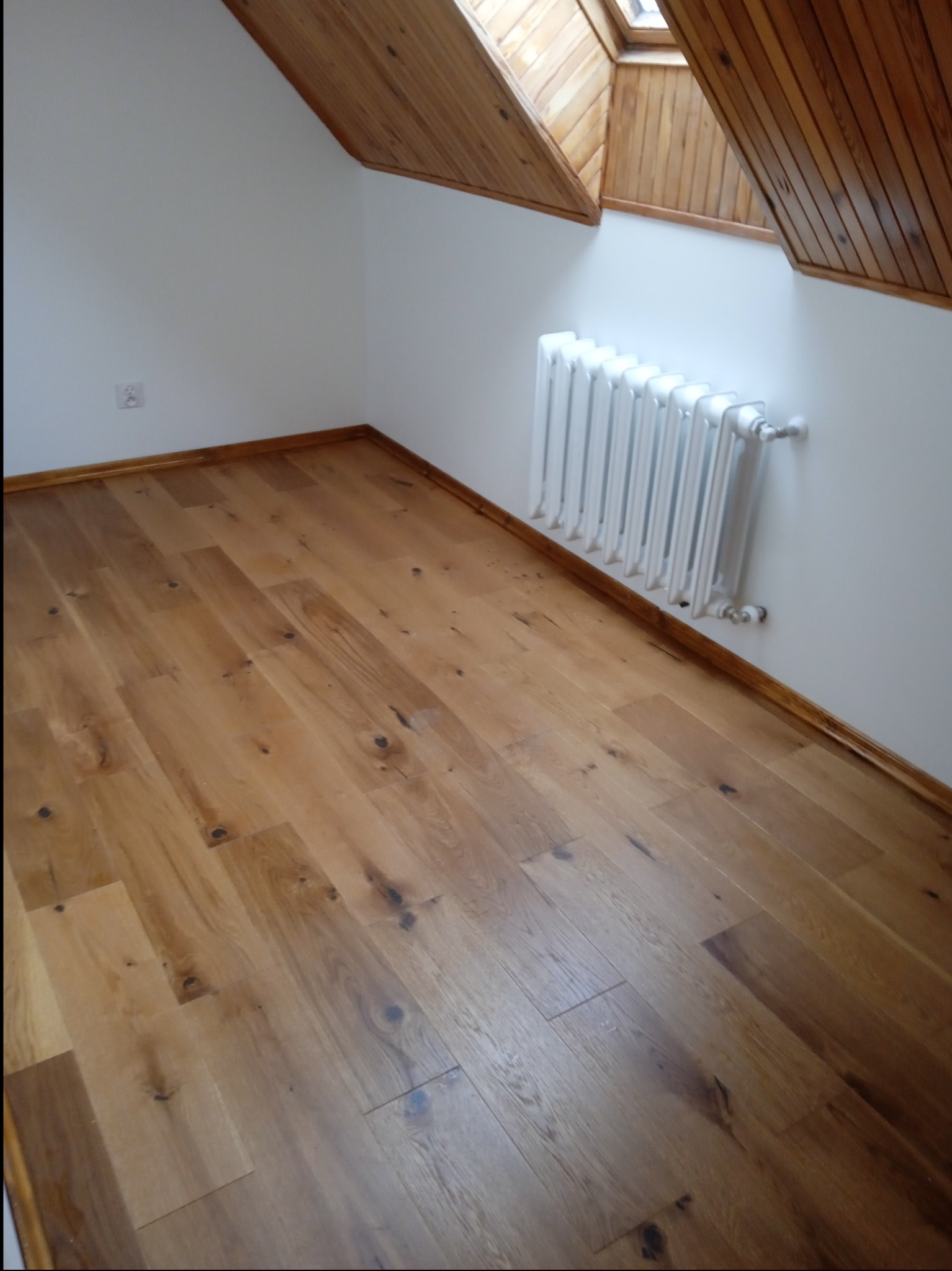 Jasne, drewniane panele podłogowe ułożone w jodełkę w pokoju na poddaszu z białymi ścianami, drewnianą boazerią na skosach dachu i białym grzejnikiem żeliwnym.