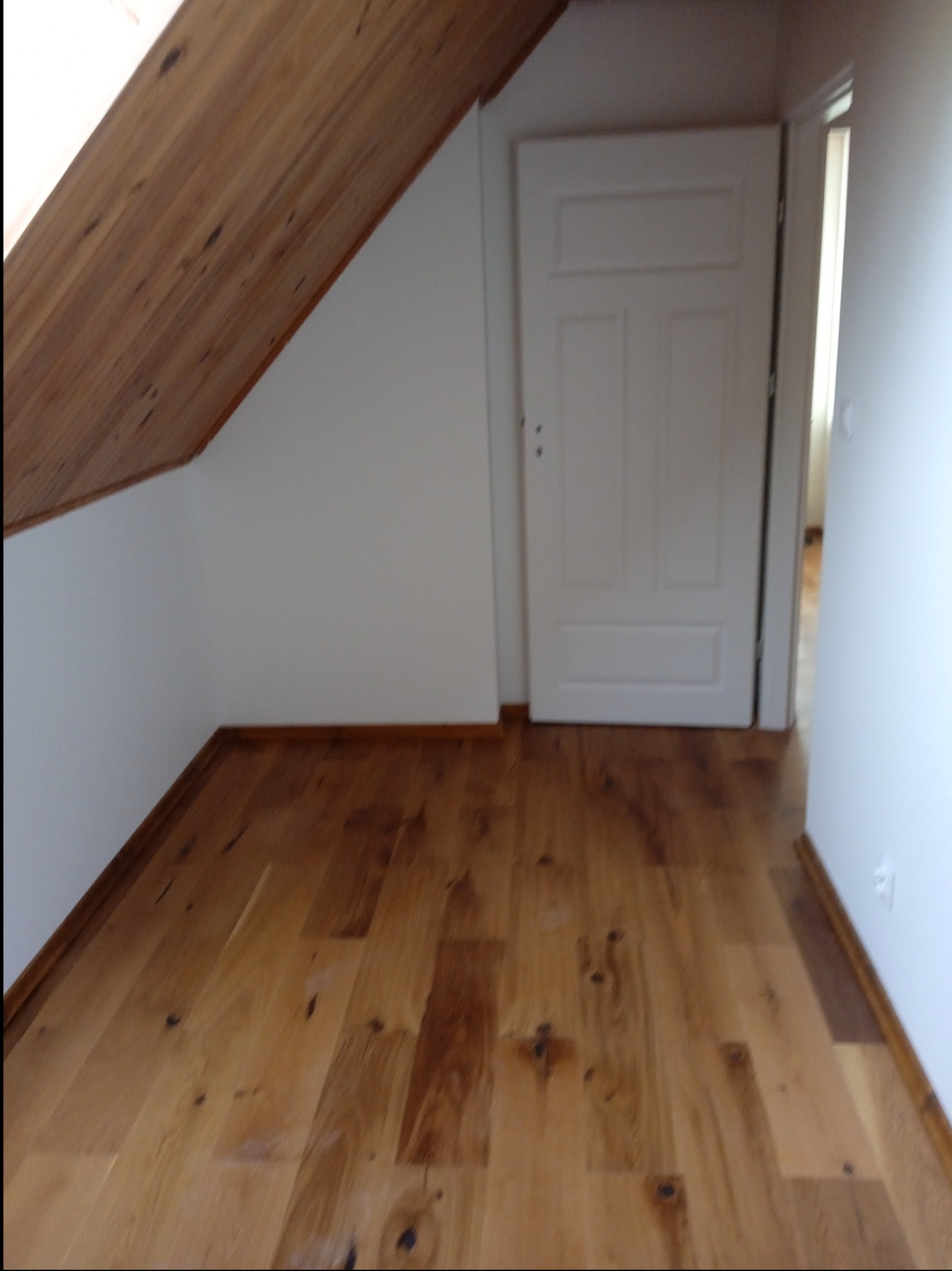 Poddasze z drewnianą podłogą i skośnym sufitem, białe ściany, białe drzwi z widocznym przejściem do innego pomieszczenia.