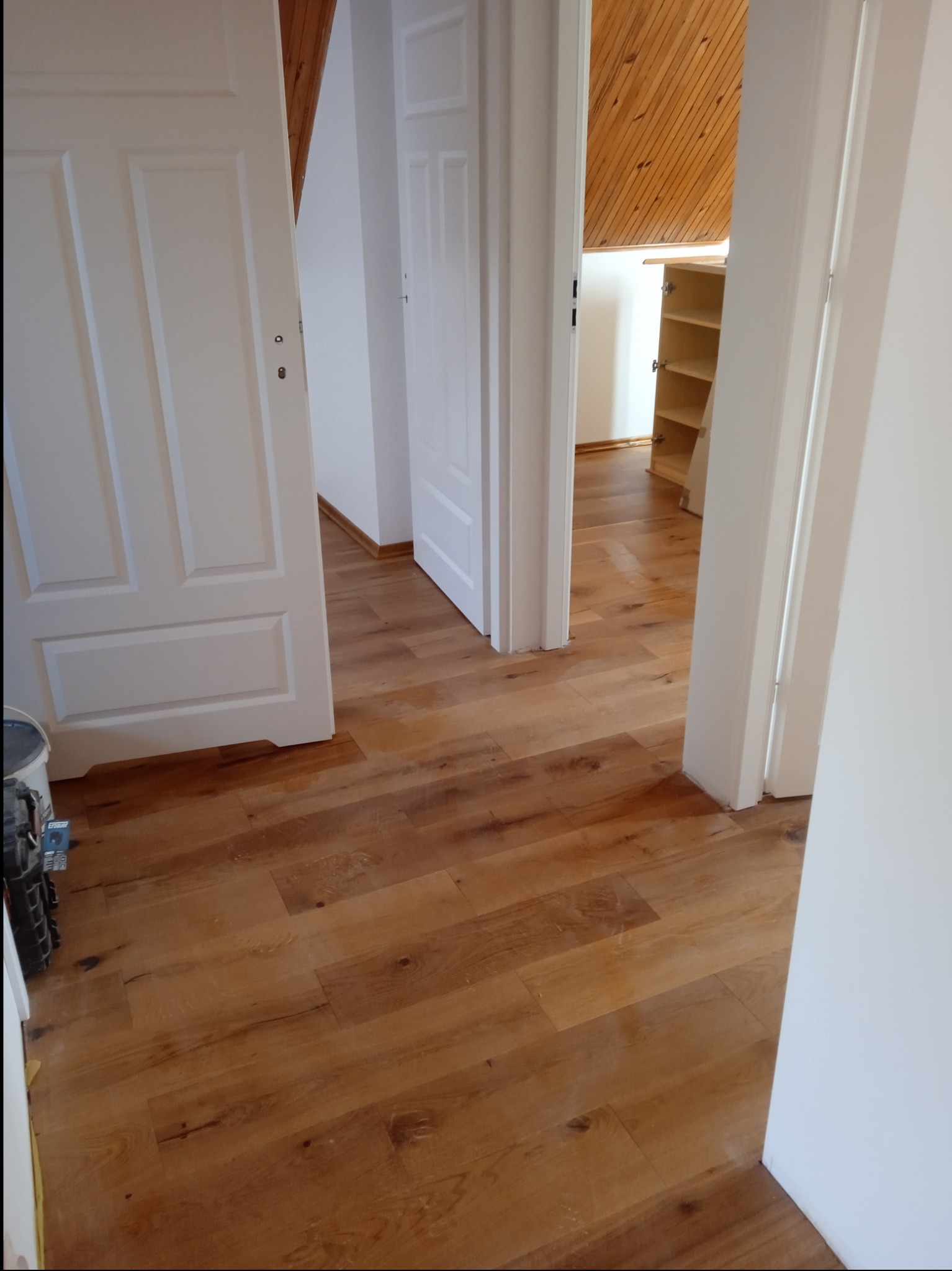 Widok na drewnianą podłogę w przejściu między pokojami, z otwartymi białymi drzwiami po obu stronach, w tle widoczna drewniana boazeria na suficie i regał.