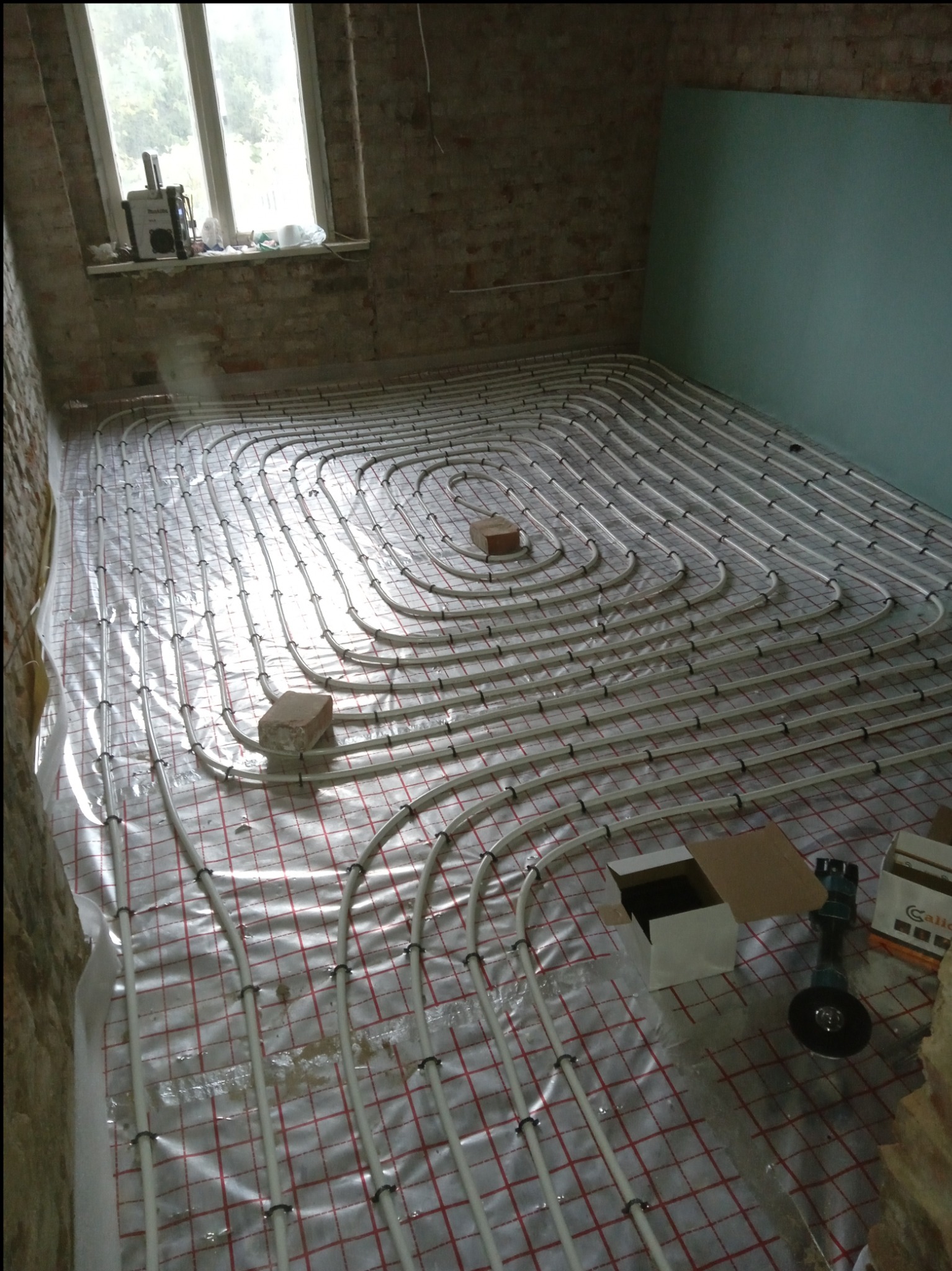 Montaż ogrzewania podłogowego w trakcie remontu: białe rurki ułożone spiralnie na folii z czerwoną kratką, przymocowane czarnymi uchwytami, widoczne ceglane ściany i narzędzia.
