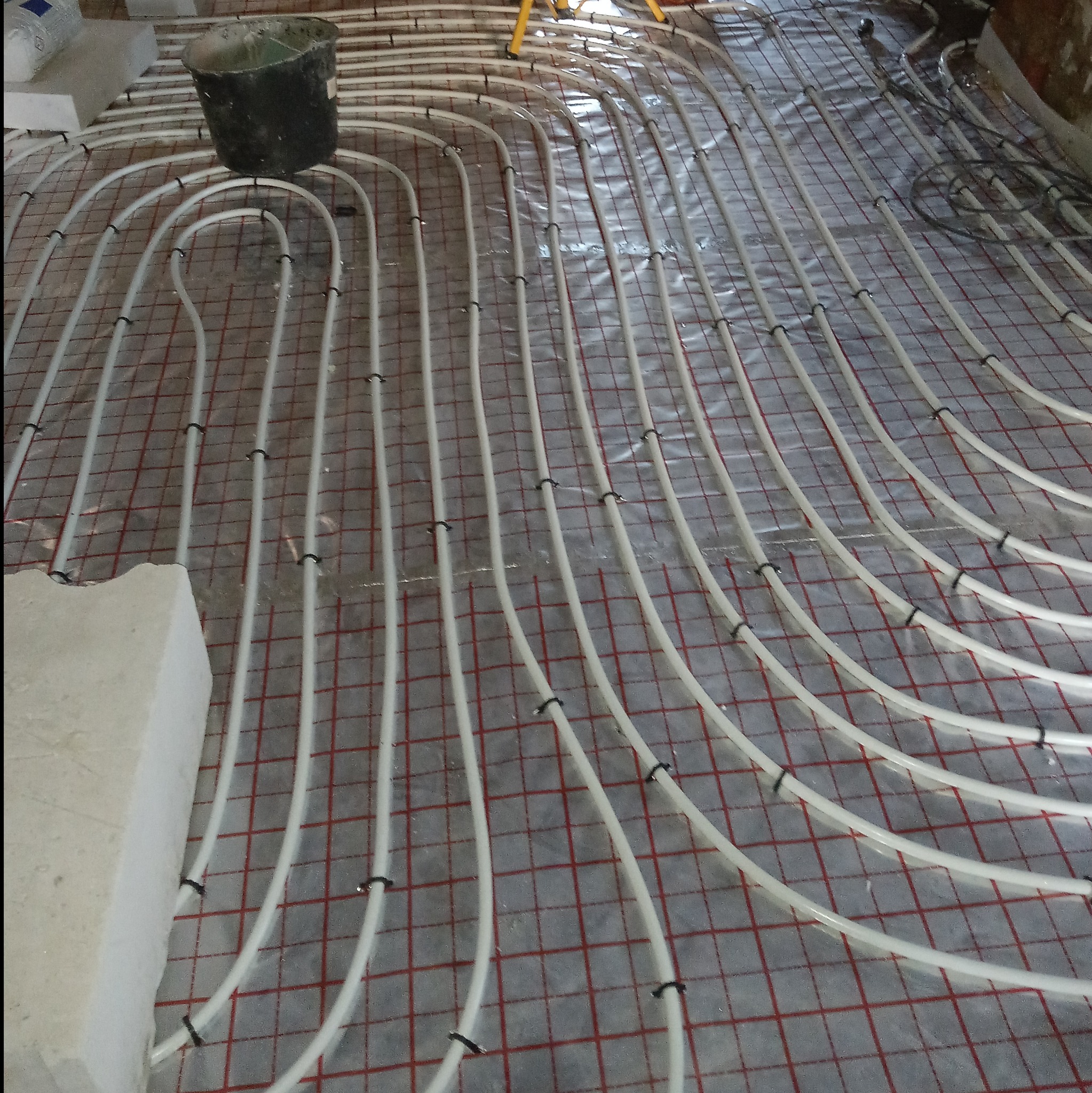 Rozłożone rury ogrzewania podłogowego na folii z siatką, widoczny kubeł i fragment żółtego statywu w tle.