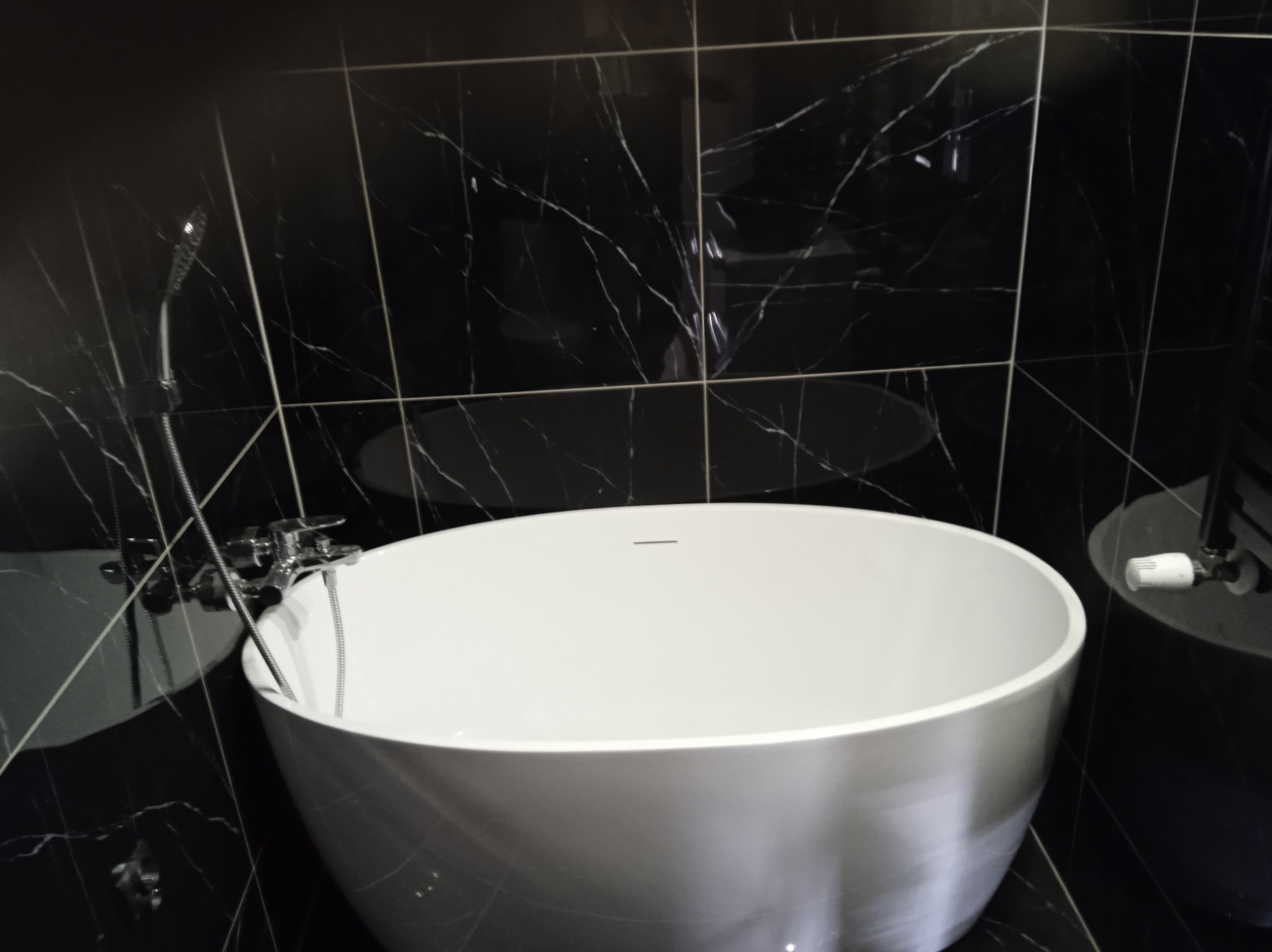 Wolnostojąca, biała wanna owalna na tle czarnych płytek imitujących marmur, chromowana bateria wannowa z prysznicem.