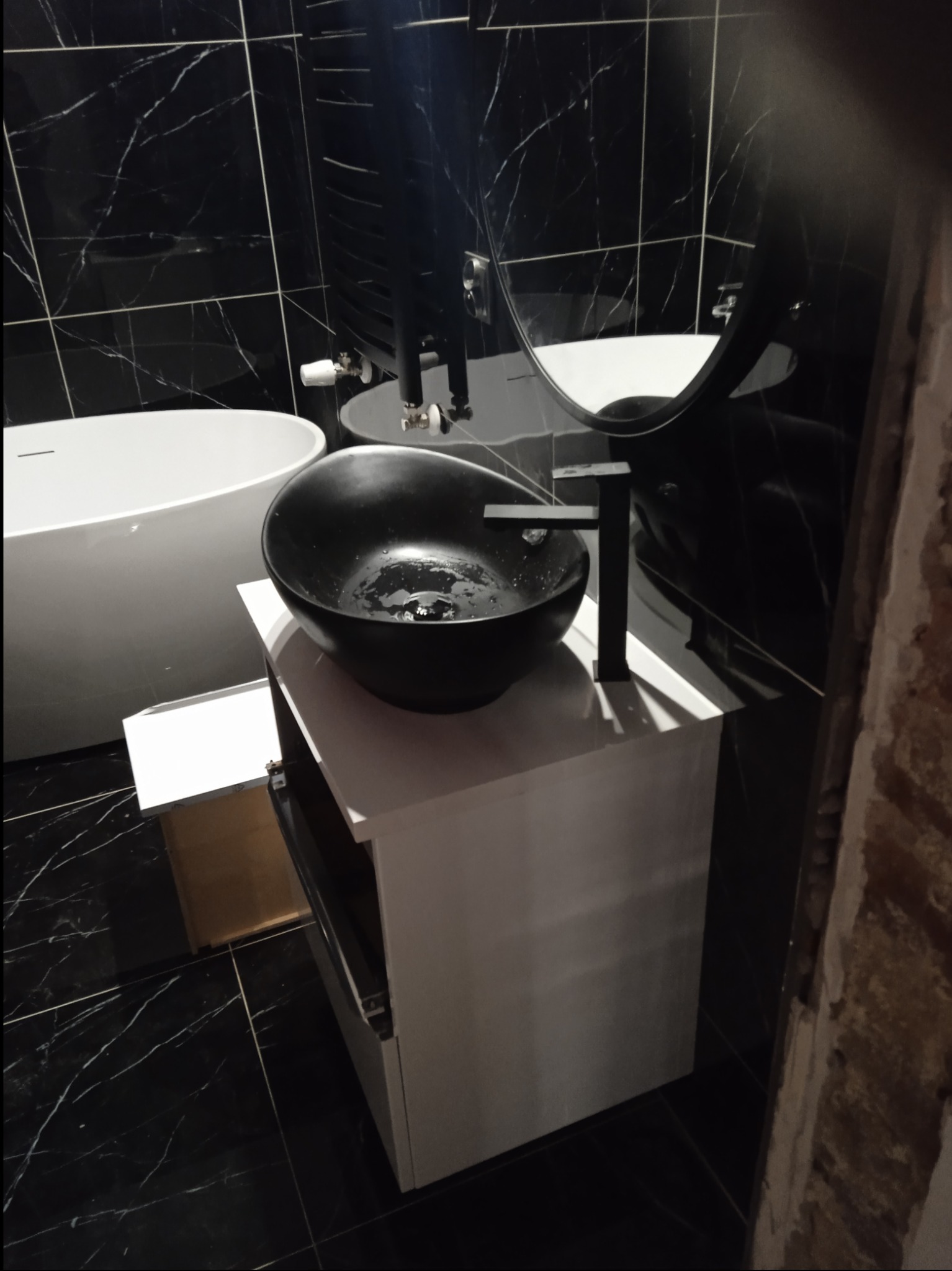 Nowoczesna łazienka z czarnymi marmurowymi płytkami, białą wolnostojącą wanną i czarną umywalką nablatową na białej szafce z otwartą szufladą.