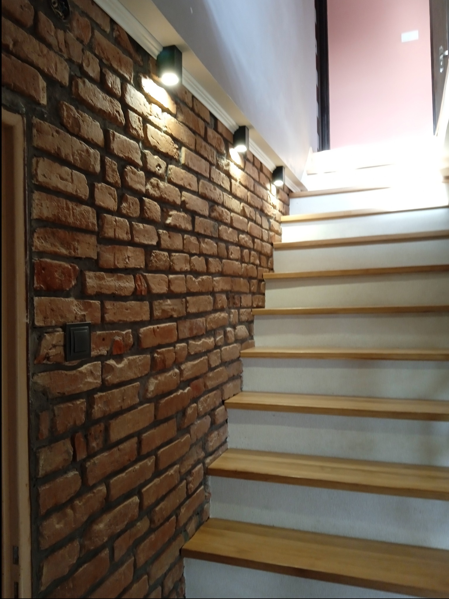 Wnętrze z ceglaną ścianą oświetloną punktowymi lampami i drewnianymi schodami z białymi podstopnicami.