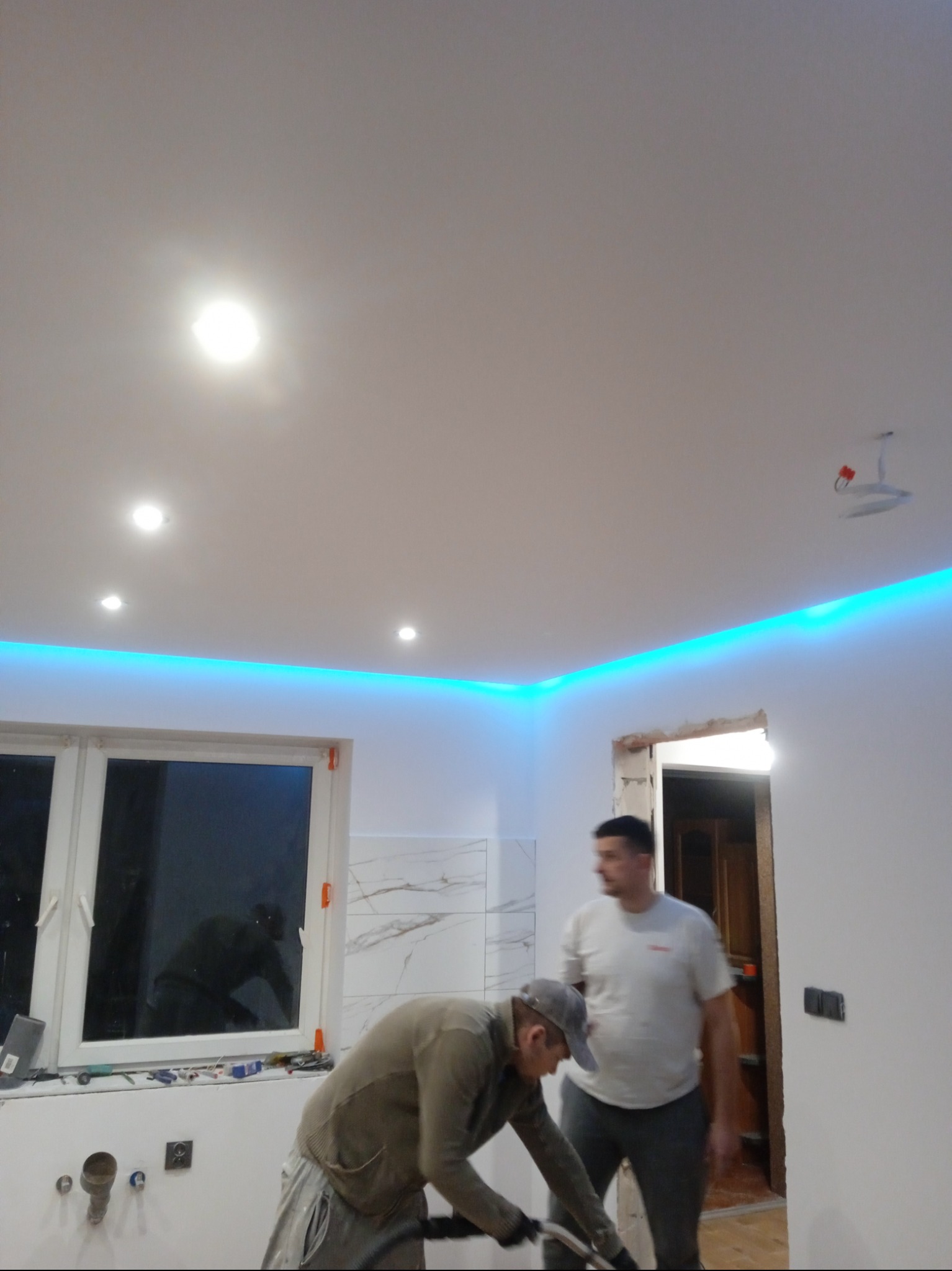 Prace wykończeniowe w pomieszczeniu: szlifowanie ściany przy oknie, widoczne oświetlenie LED podsufitowe, fragment marmurowej okładziny ściennej i mężczyzna w tle.
