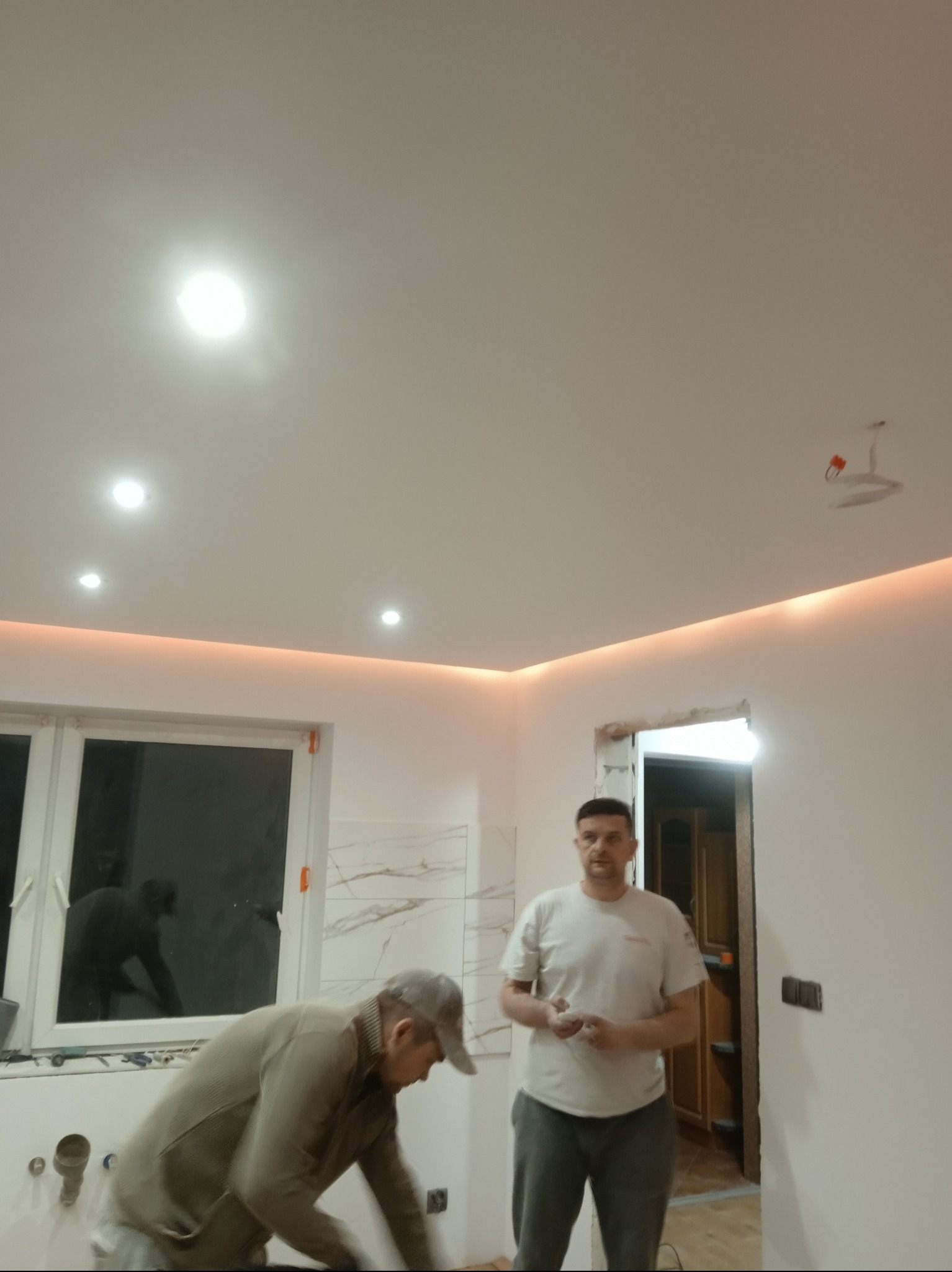 Dwaj mężczyźni w trakcie prac wykończeniowych w pomieszczeniu z oświetleniem LED podsufitowym i płytkami imitującymi marmur, jeden z nich pracuje przy oknie, drugi stoi w pobliżu otwartych drzwi.