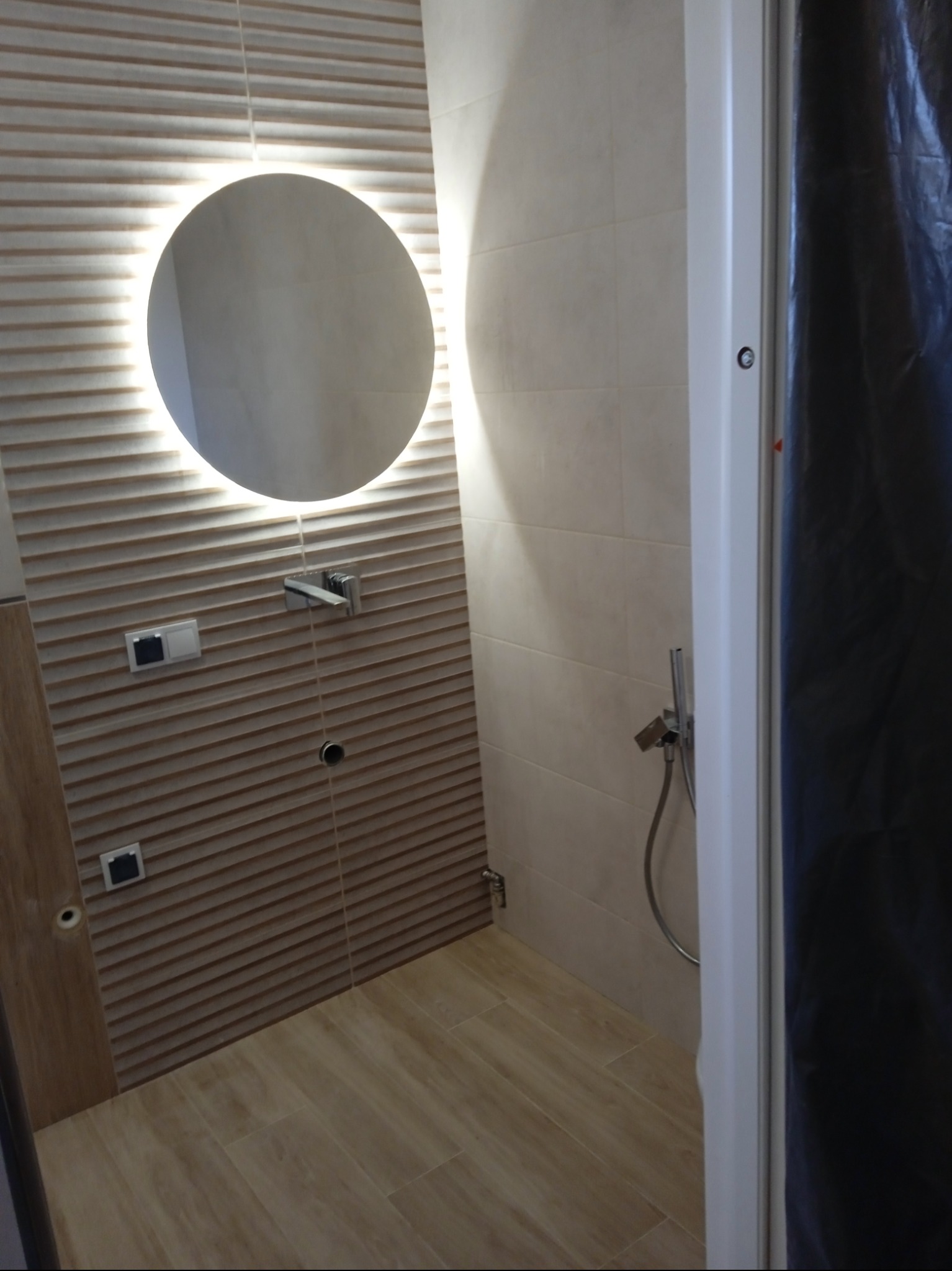 Nowoczesna łazienka w Olecku z okrągłym lustrem podświetlanym LED, beżowe płytki imitujące drewno na ścianach i podłodze, chromowana armatura i częściowo zasłonięta kabina prysznicowa.