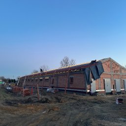 NORD INVEST - Budynek gospodarczy w trakcie budowy dachu, widoczni dekarze pracujący na wysokości, ceglana konstrukcja ścian, teren budowy z ziemią i materiałami budowlanymi, tablica z nazwą miejscowości w tle.