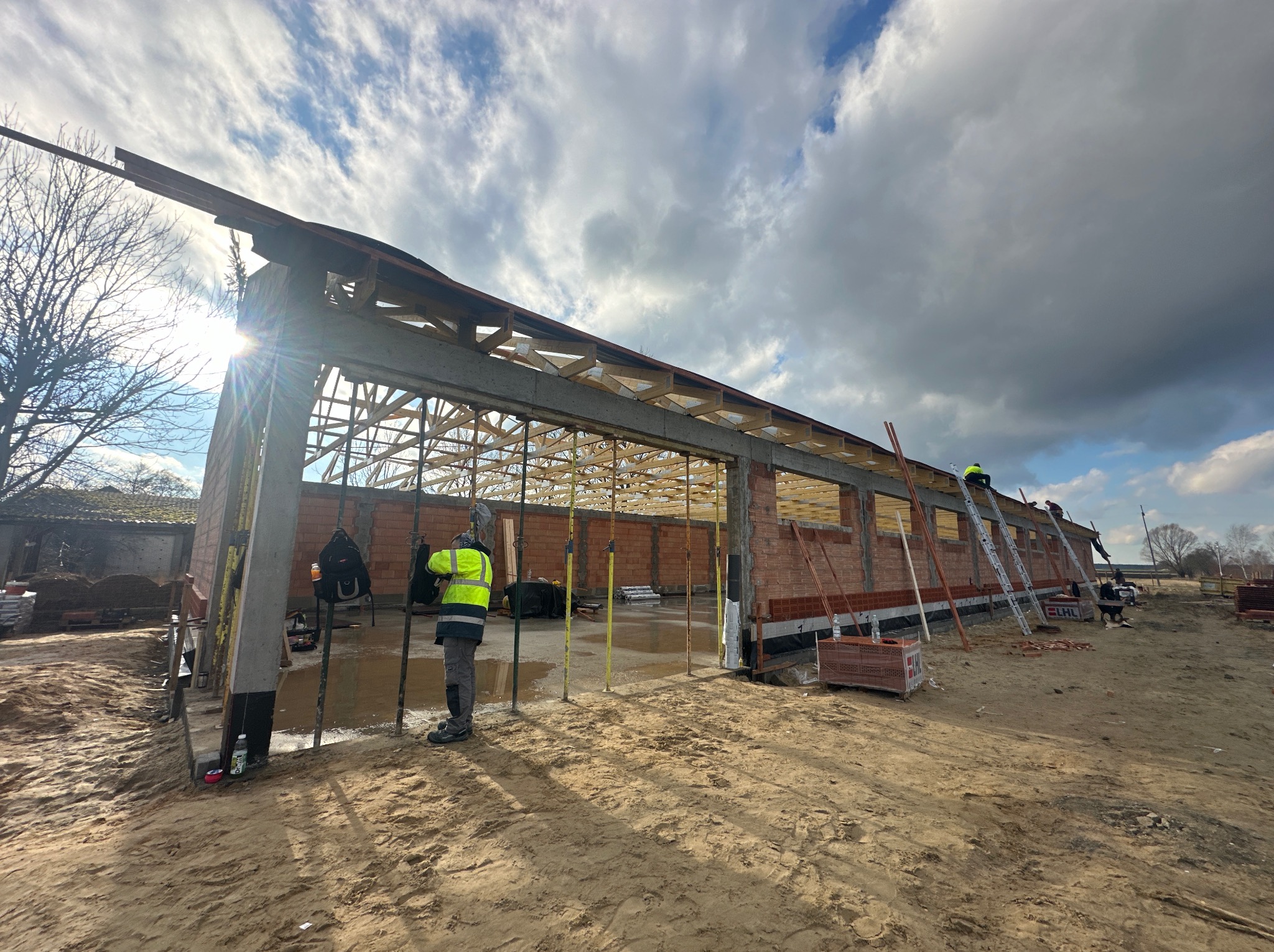 Budowa domu w stanie surowym otwartym: murarz w żółtej kamizelce ustawia podpory stropowe, w tle widać szkielet dachu i pochmurne niebo.