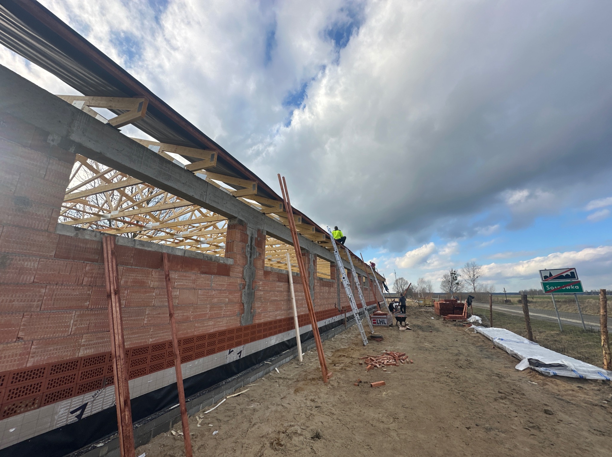 Budowa domu jednorodzinnego w stanie surowym otwartym: widoczna konstrukcja dachu z drewnianą więźbą, ściany z czerwonej cegły, pracownicy na drabinach i materiały budowlane na placu budowy...