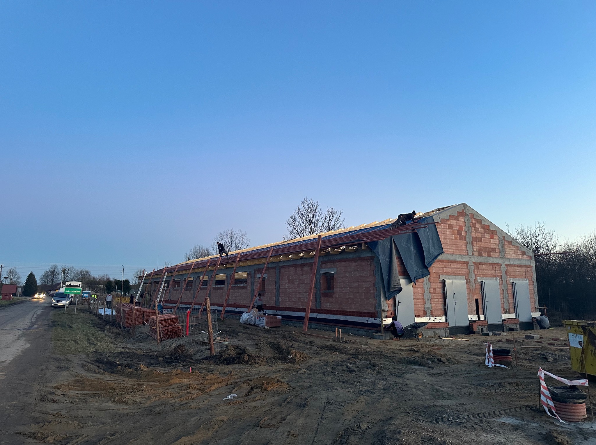 Budynek gospodarczy w trakcie budowy dachu, widoczni dekarze pracujący na wysokości, ceglana konstrukcja ścian, teren budowy z ziemią i materiałami budowlanymi, tablica z nazwą miejscowości w tle.