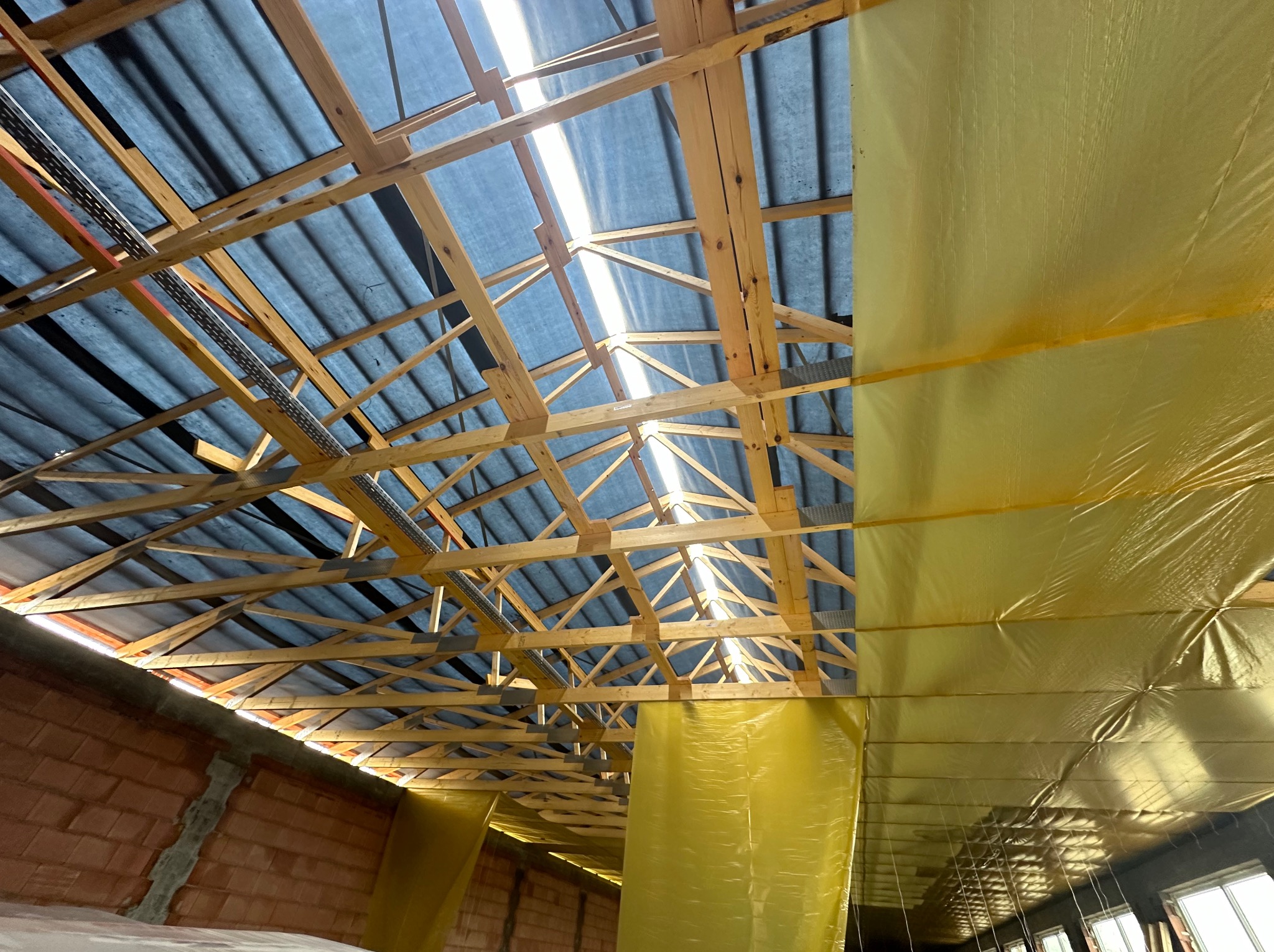 Wnętrze dachu w trakcie krycia, widoczna drewniana konstrukcja, folia paroizolacyjna w kolorze żółtym i prześwitujące niebieskie pokrycie dachowe z widocznym światłem słonecznym.