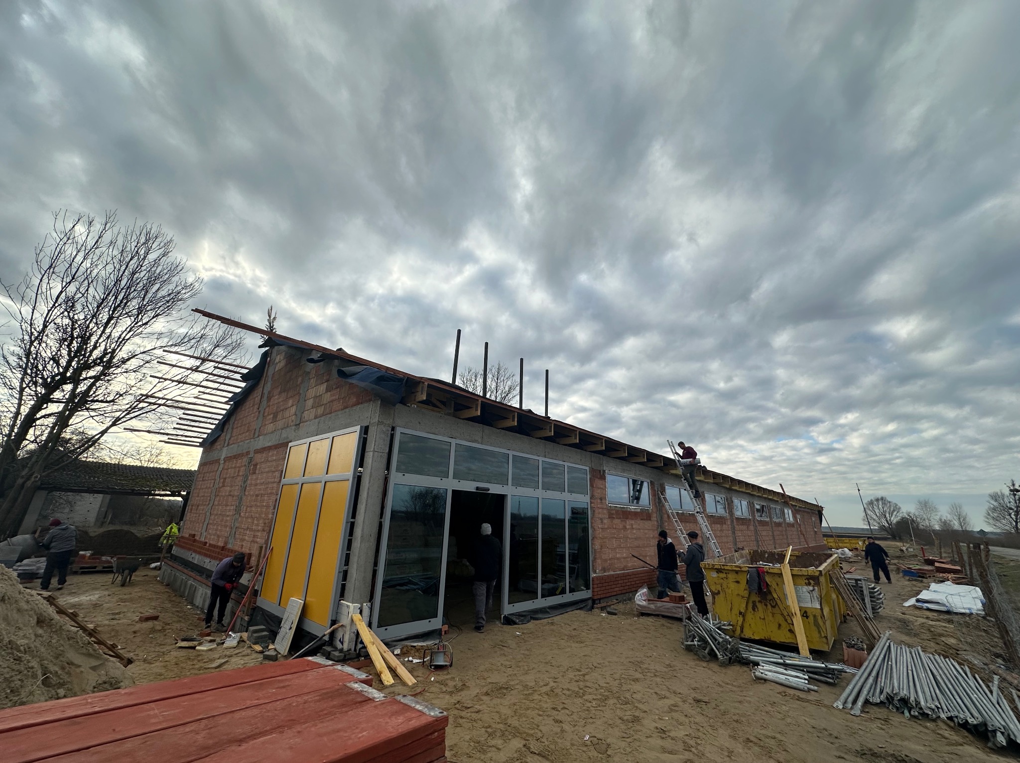 Budynek w trakcie prac dekarskich i montażu stolarki okiennej; widoczni pracownicy, rusztowania, kontener na odpady budowlane i częściowo zdjęte poszycie dachu przy pochmurnym niebie.