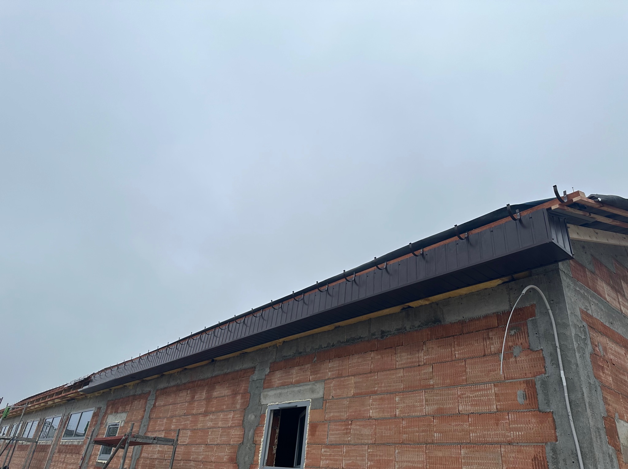 Budynek w trakcie budowy w Poznaniu z zamontowaną, brązową podbitką dachową i orynnowaniem, widoczne cegły i szare niebo.