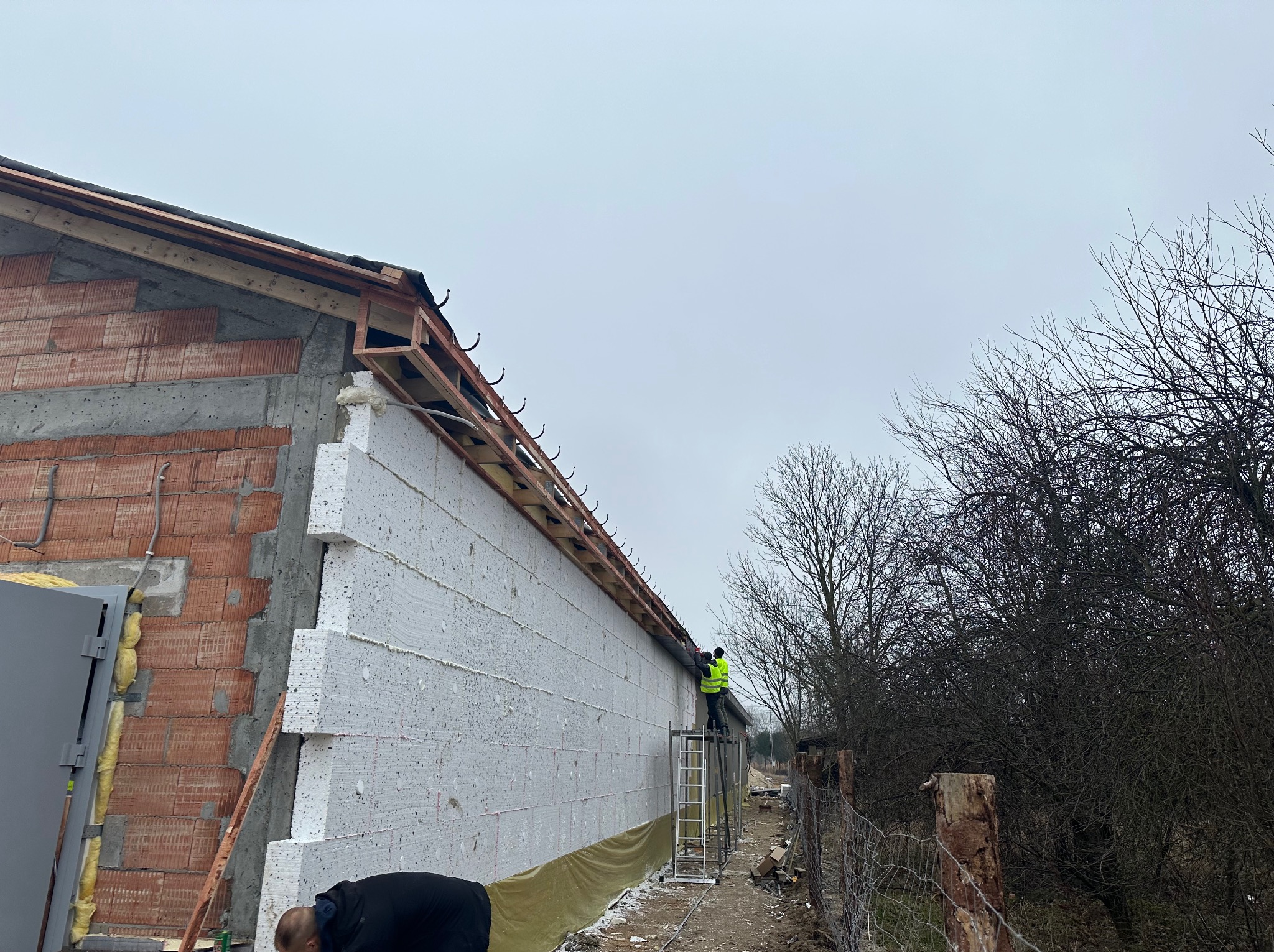 Montaż ocieplenia budynku z pustaków ceramicznych styropianem, widoczni dwaj pracownicy na drabinie w żółtych kamizelkach, przygotowujący konstrukcję pod dach.