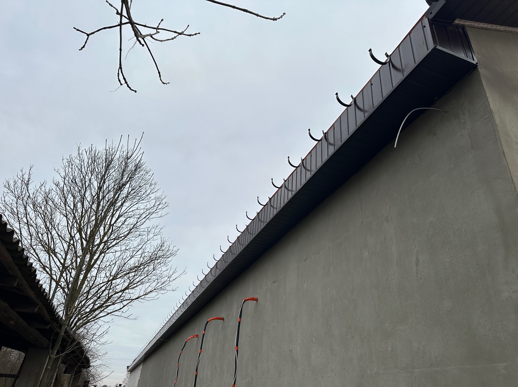 Nowe orynnowanie w kolorze ciemnego brązu zamontowane na świeżo otynkowanej ścianie budynku, widoczne uchwyty rynnowe i trzy rury spustowe z pomarańczowymi końcówkami, widok z dołu na tle...