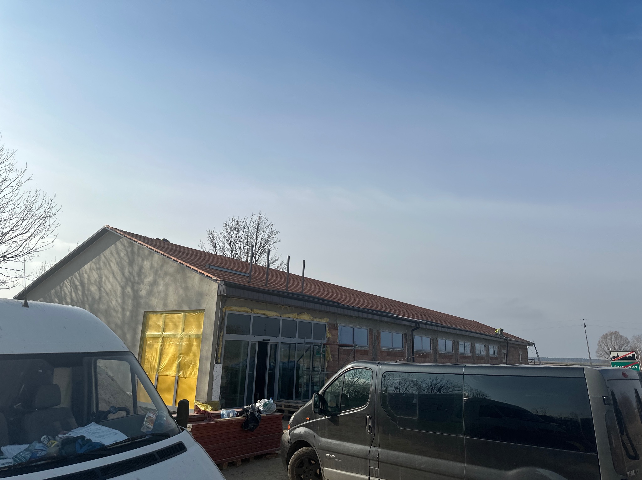 Budynek w trakcie remontu z częściowo wymienionym dachem, widoczne dwie furgonetki na pierwszym planie i materiały budowlane składowane obok budynku.