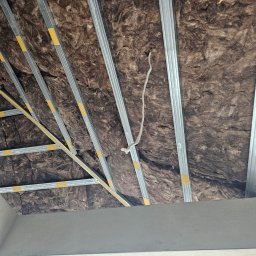 MAX-BUD JAKUB ZARZYŃSKI - Ocieplenie poddasza wełną mineralną widoczne pomiędzy metalowymi profilami, z wystającym kablem elektrycznym.