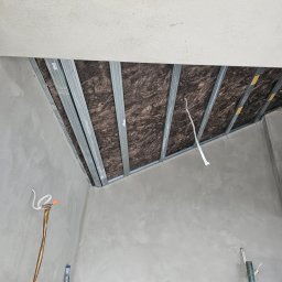 MAX-BUD JAKUB ZARZYŃSKI - Ocieplenie poddasza wełną mineralną, widoczne metalowe profile konstrukcyjne, surowe ściany i wystające przewody elektryczne.