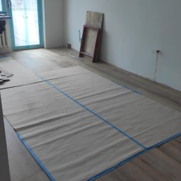 MAX-BUD JAKUB ZARZYŃSKI - Przygotowanie do malowania: podłoga zabezpieczona płachtą malarską z niebieską taśmą. Widoczne panele podłogowe, okno z folią ochronną i fragment ściany.