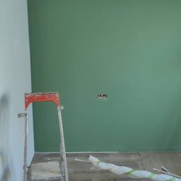 MAX-BUD JAKUB ZARZYŃSKI - Ściana pomalowana na zielono z widoczną drabiną malarską i akcesoriami malarskimi. Przygotowanie do dalszych prac wykończeniowych, zabezpieczona listwa sufitowa.