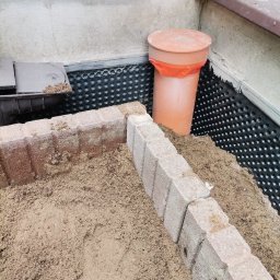 MAX-BUD JAKUB ZARZYŃSKI - Fundament izolowany folią kubełkową, obłożony cegłą klinkierową i zasypany piaskiem, z widoczną rurą kanalizacyjną w rogu. Prace ziemne.