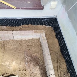 MAX-BUD JAKUB ZARZYŃSKI - Fundament zabezpieczony folią kubełkową i obłożony cegłą. Etap prac związany z izolacją przeciwwilgociową fundamentów budynku. Widoczny fragment ściany i okna.