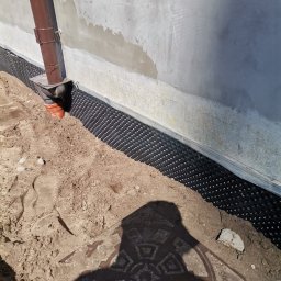 MAX-BUD JAKUB ZARZYŃSKI - Izolacja fundamentu budynku z folią kubełkową i rurą spustową, widoczne ślady świeżego tynku. Prace ziemne w toku, widoczny właz kanalizacyjny.