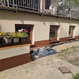 MAX-BUD JAKUB ZARZYŃSKI - Dwukolorowa elewacja domu z oknami, kwiatami w donicach i zabezpieczeniem fundamentów folią. Widoczny fragment balkonu z metalową barierką.