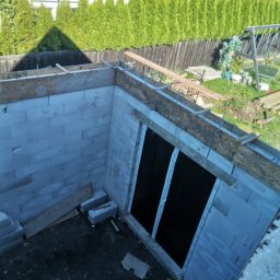 MAX-BUD JAKUB ZARZYŃSKI - Wznoszenie ścian fundamentowych z bloczków betonowych, widok z góry na budowę, zbrojenie i szalunki, zielone tuje w tle.