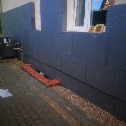 MAX-BUD JAKUB ZARZYŃSKI - Ocieplenie fundamentów budynku płytami styropianowymi w kolorze grafitowym, widoczne okno z odbiciem nieba i fragment dachu sąsiedniego domu.