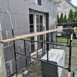 Ocieplanie domu szarym styropianem grafitowym. Widoczne rusztowanie i okno. Prace w toku, pianka montażowa uszczelnia łączenia płyt. Dom jednorodzinny w tle.