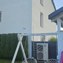 MAX-BUD JAKUB ZARZYŃSKI - Biały, dwupiętrowy dom z klimatyzacją na zewnątrz, widziany z ogrodu z huśtawką i zjeżdżalnią. Świeżo pomalowana elewacja w słoneczny dzień.