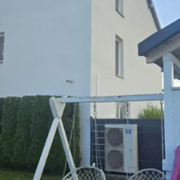 Biały, dwupiętrowy dom z klimatyzacją na zewnątrz, widziany z ogrodu z huśtawką i zjeżdżalnią. Świeżo pomalowana elewacja w słoneczny dzień.