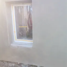 Świeżo położona, szara gładź na elewacji domu, okno zabezpieczone folią i taśmą malarską. Widoczna siatka na ościeżnicy okiennej.