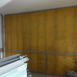 MAX-BUD JAKUB ZARZYŃSKI - Ocieplenie ściany wełną mineralną w metalowym stelażu, widoczne płyty gipsowe i narzędzia. Etap prac wykończeniowych wewnątrz pomieszczenia.