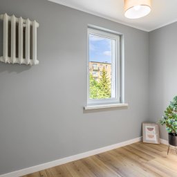 MAX-BUD JAKUB ZARZYŃSKI - Szare wnętrze pokoju po malowaniu ścian z białym grzejnikiem, oknem z widokiem na budynek i drzewa, drewnianą podłogą oraz rośliną doniczkową w rogu.
