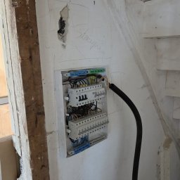 MAX-BUD JAKUB ZARZYŃSKI - Rozdzielnica elektryczna zamontowana na ścianie w trakcie prac remontowych, widoczne kable i elementy instalacji w surowym wnętrzu.