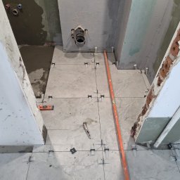 MAX-BUD JAKUB ZARZYŃSKI - Układanie szarych płytek podłogowych w łazience, widoczne poziomica, krzyżyki dystansowe i fragment instalacji kanalizacyjnej. Etap prac wykończeniowych.