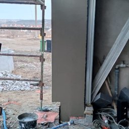MAX-BUD JAKUB ZARZYŃSKI - Świeżo otynkowany filar budynku w trakcie prac wykończeniowych, widoczne rusztowanie, narzędzia i wiadro z zaprawą. Tło stanowią zabudowania w oddali.