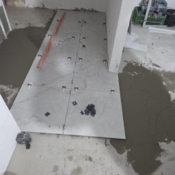 MAX-BUD JAKUB ZARZYŃSKI - Układanie płytek podłogowych w trakcie remontu. Widoczne płytki, poziomica, krzyżyki dystansowe i zaprawa. Fragment podłogi przed wykończeniem.