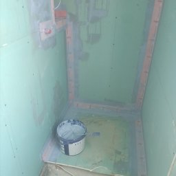 MAX-BUD JAKUB ZARZYŃSKI - Przygotowanie do malowania w łazience: ściany z płyt gipsowych, zabezpieczone narożniki, wiadro z farbą i pędzel na podłodze. Widok z góry na wnętrze w trakcie prac.