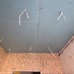 MAX-BUD JAKUB ZARZYŃSKI - Montaż płyt gipsowo-kartonowych na suficie, widoczne kable i surowe ściany z płyty OSB i betonu. Etap prac wykończeniowych w pomieszczeniu.