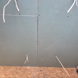 MAX-BUD JAKUB ZARZYŃSKI - Płyty gipsowo-kartonowe na suficie z wystającymi przewodami elektrycznymi, widoczne łączenia płyt i fragment ściany z surowego betonu oraz płyty OSB na ścianie.