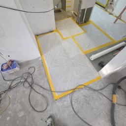 MAX-BUD JAKUB ZARZYŃSKI - Przygotowanie podłoża: zabezpieczona folią i taśmą podłoga w trakcie prac wykończeniowych. Widoczne kable, narzędzia i fragment buta pracownika.